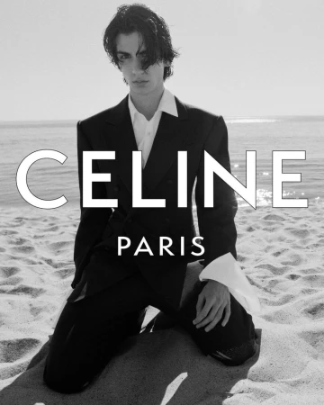 Male model kneeling on a sandy beach wearing a dark tailored suit jacket and white collared shirt.