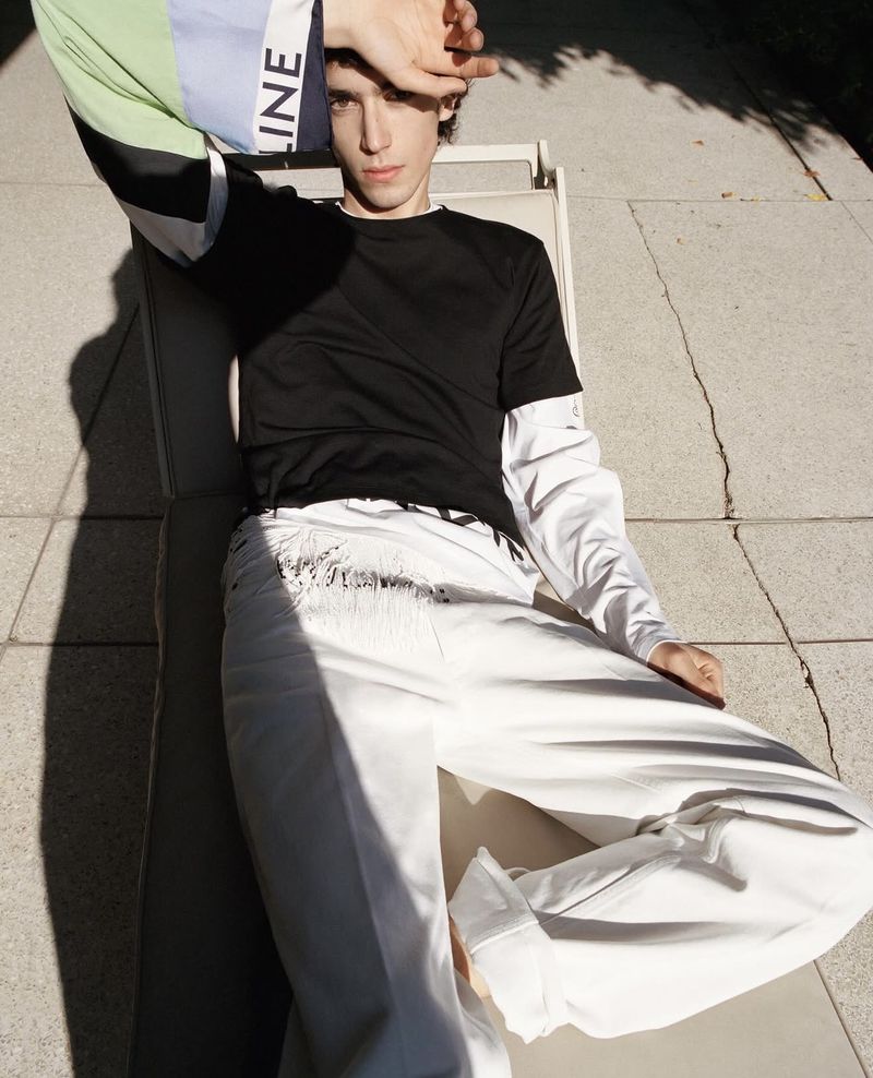 A male model reclining on an outdoor lounge chair in bright sunlight wearing a black t-shirt and wide-leg white trousers.