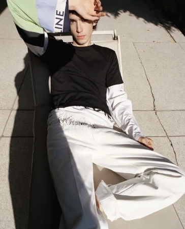 A male model reclining on an outdoor lounge chair in bright sunlight wearing a black t-shirt and wide-leg white trousers.