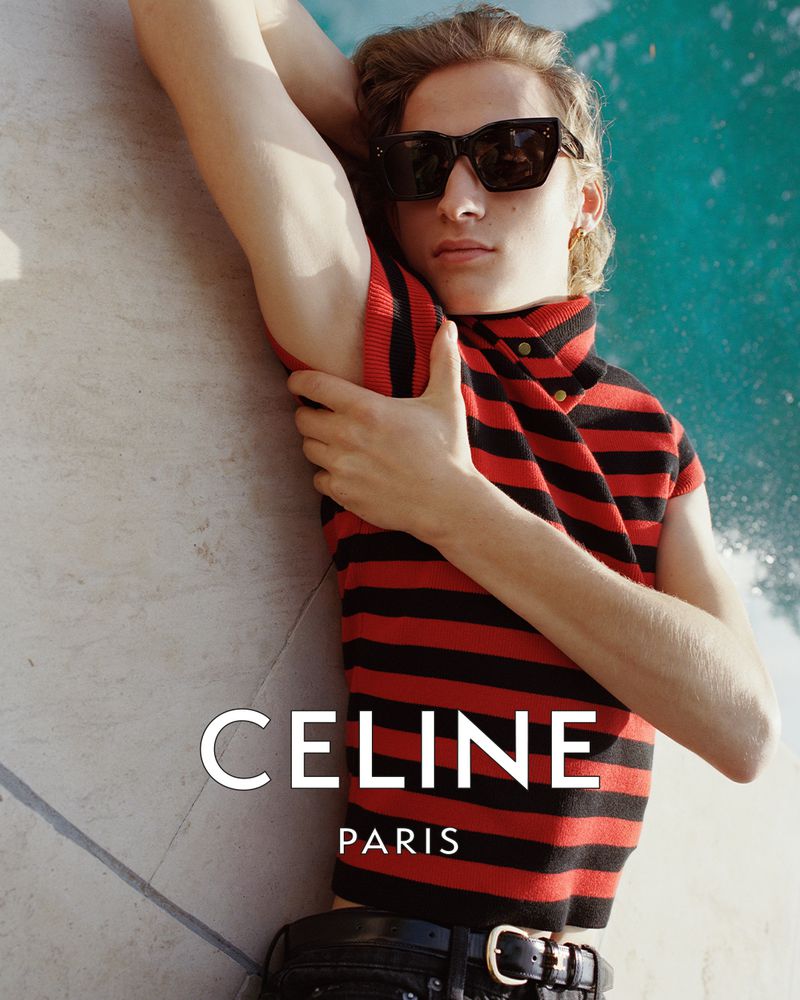Model wearing dark sunglasses and a red and black striped sleeveless top reclining poolside next to clear turquoise water.