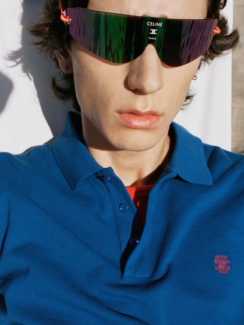 Close-up portrait of a male model wearing iridescent Celine shield sunglasses and a bright blue polo shirt in direct sunlight.
