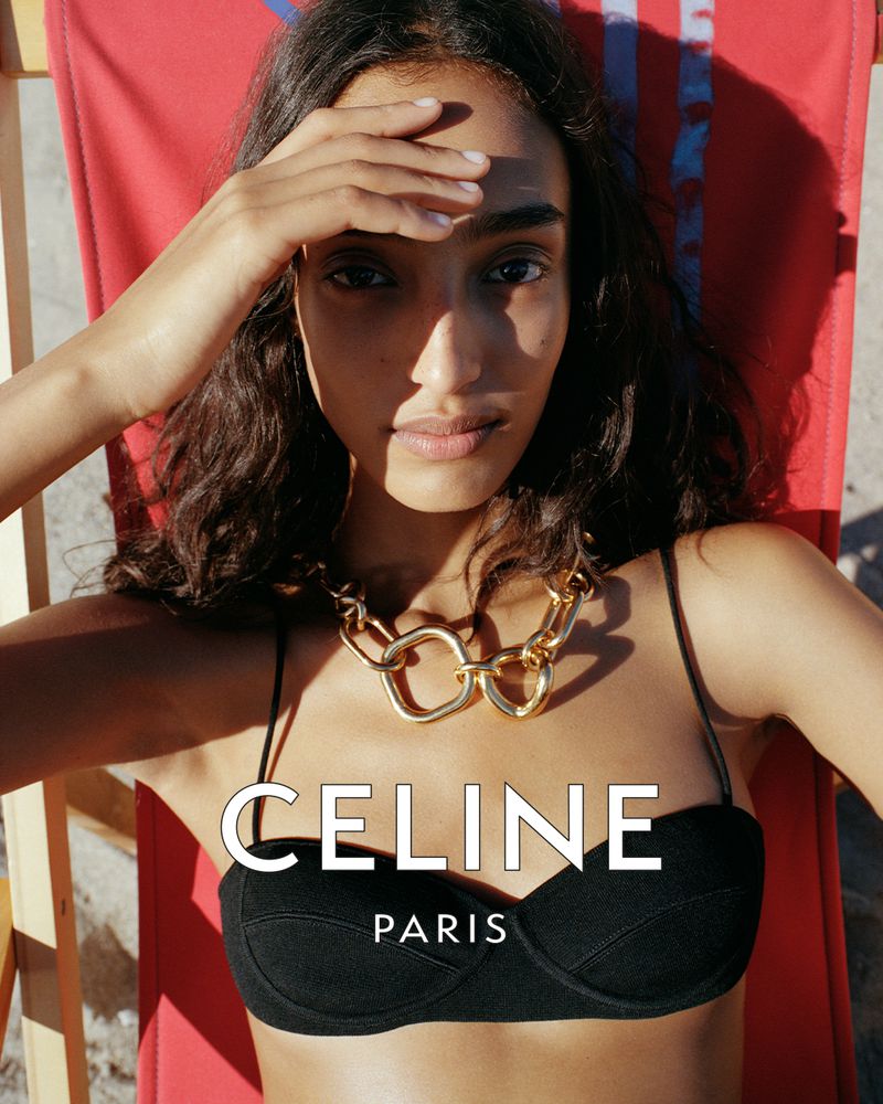 Model Mona Tougaard wearing a black bikini top and a chunky gold necklace while reclining on a red beach chair for Celine.