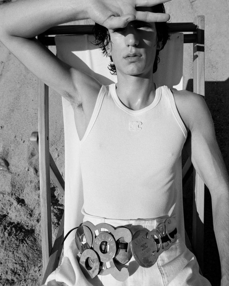Black and white photo of a man reclining on a canvas beach chair wearing a white ribbed tank top and a wide metallic emblem belt.