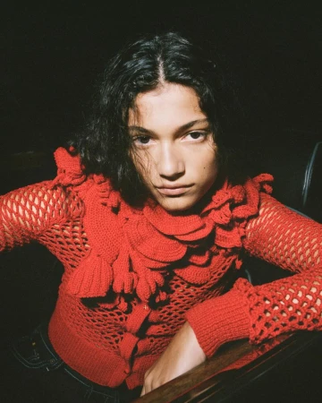 A model wearing an open-knit red ruffled sweater leans forward against a dark backdrop in a close-up portrait.