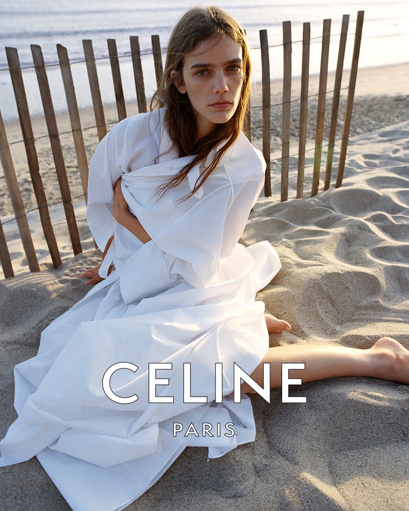 Model seated on a sandy beach wearing a voluminous white wrap dress near a wooden fence for Celine.