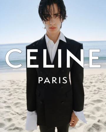 Model wearing a black tailored double-breasted blazer and white collared shirt standing on a sandy beach for Celine.