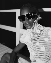 Mona Tougaard wearing black rectangular sunglasses and a white open-knit high-neck top in a high-contrast black and white portrait.