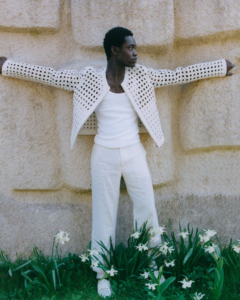A black male model leans against a textured beige wall in an open-weave jacket, tank top, and trousers, surrounded by greenery and white flowers.