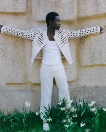 A black male model leans against a textured beige wall in an open-weave jacket, tank top, and trousers, surrounded by greenery and white flowers.