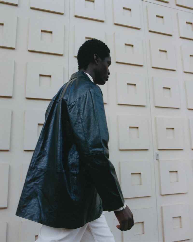 Fashionable Black male model strides outdoors in a dark jacket and white pants, posing against a cream-colored square-patterned textured wall.