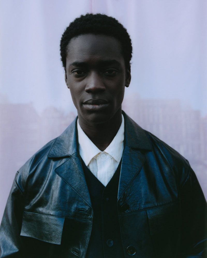 Portrait of an African-American adult male wearing a dark navy leather jacket with a white button-down shirt on a light purple background.