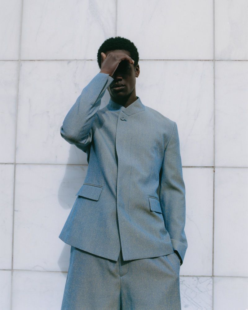 Man in light blue suit shields eyes in outdoor fashion shot against a white marble-tile wall in soft, diffused light.