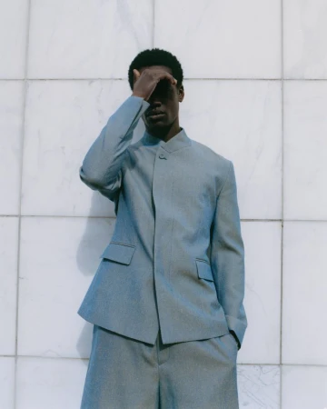 Man in light blue suit shields eyes in outdoor fashion shot against a white marble-tile wall in soft, diffused light.