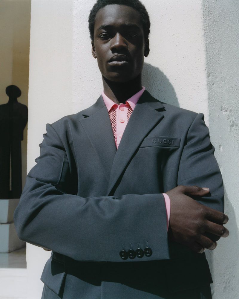 An African American man in a dark gray suit and pink shirt stands confidently in front of a white wall in a portrait-style shot.