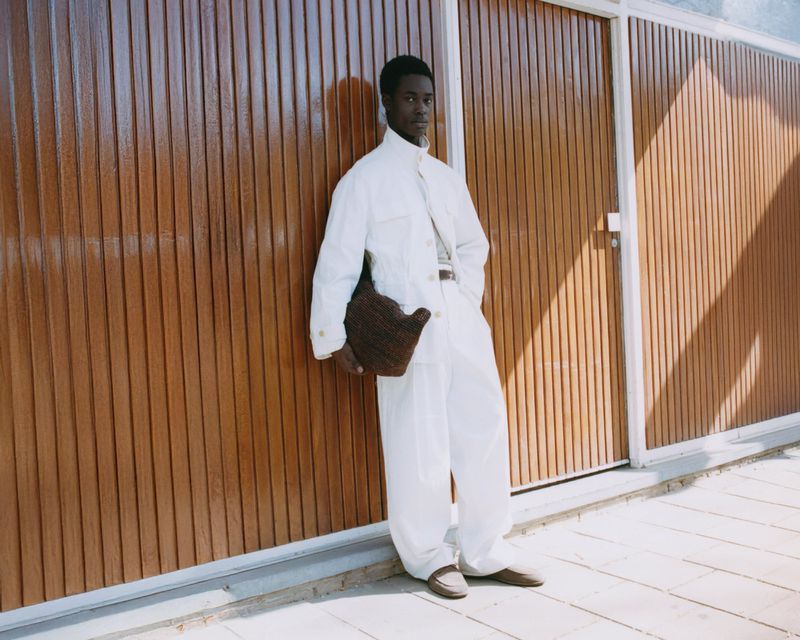 An African American man stands next to a textured brown wall wearing an off-white jumpsuit and carrying a brown woven bag.