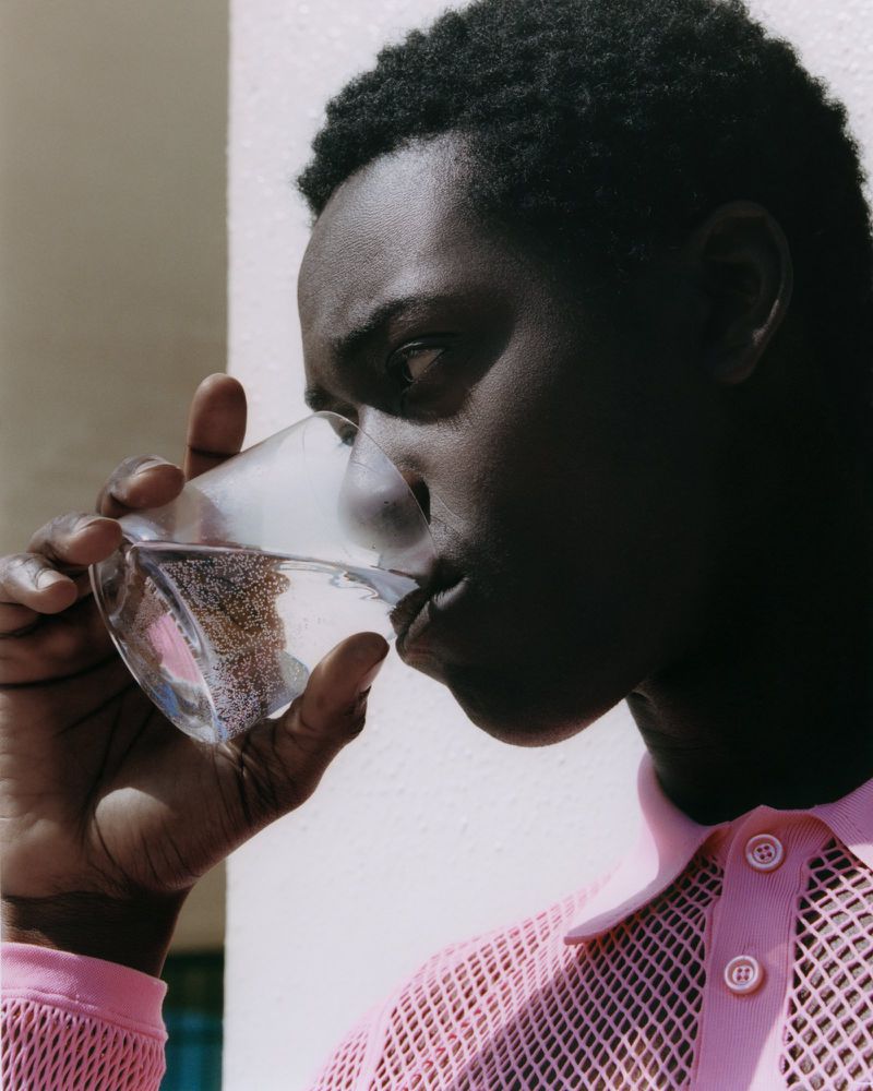 Young man drinks clear liquid from glass in bright light, wearing pink mesh shirt, close-up portrait orientation.