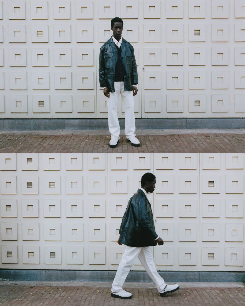 Dark-skinned man in an overshirt, sweater, and white pants poses against a textured wall in full and side profile views, outdoors.
