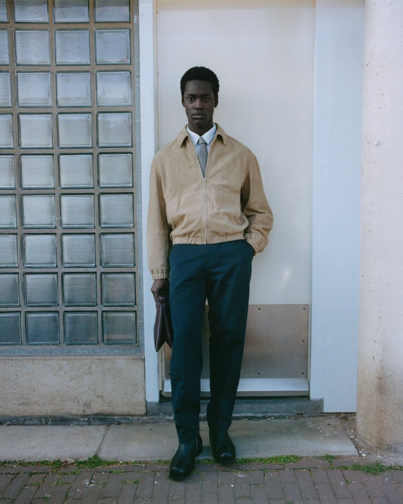 Dark-skinned adult male poses against wall wearing tan jacket, shirt, tie, and dark slacks, holding dark leather document case.