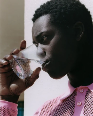 Profile portrait of model Yaw Tiëku Appiah drinking water from a clear glass, wearing a light pink collared mesh shirt.