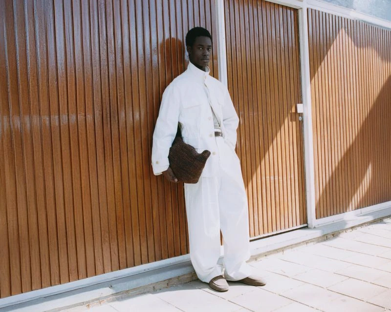 Model Yaw Tieku Appiah in a white jacket and trousers holding a brown bag against a slatted wooden wall for The Greatest Magazine.