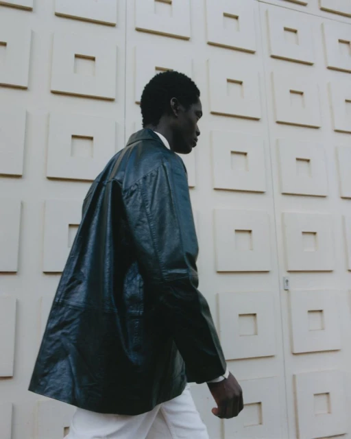 Profile view of a black man wearing a dark leather jacket and white trousers against a patterned architectural wall.