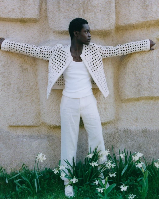 Model Yaw Tieku Appiah posing against a textured stone wall wearing a white open-weave jacket and light trousers for The Greatest Magazine.