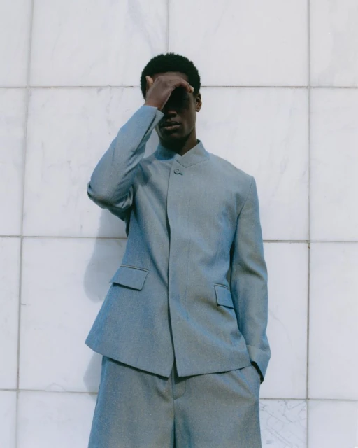 Model Yaw Tiëku Appiah wearing a light blue tailored stand-collar suit while shading his face against a marble tiled wall.
