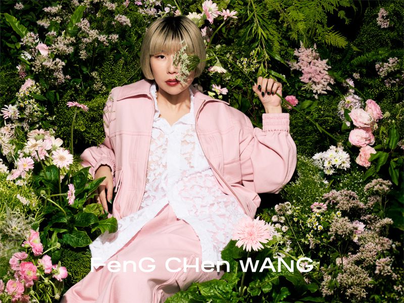 East Asian woman with a blonde bob wearing a pink jacket and lace shirt, seated amid dense foliage and pink flowers.