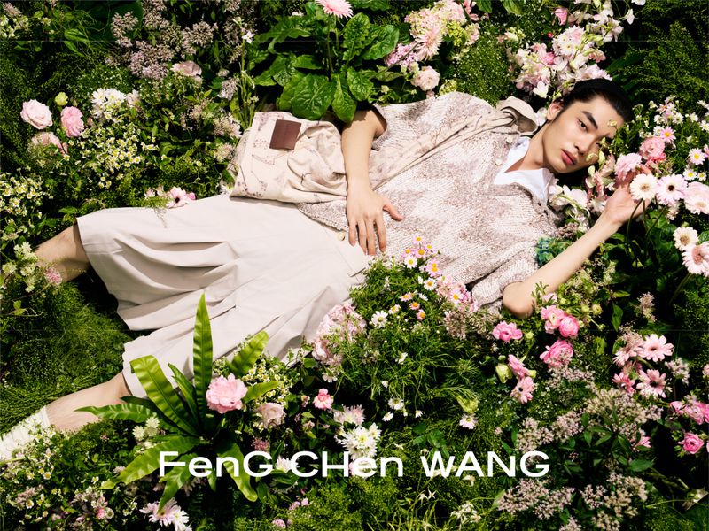 An East Asian male model lying among lush greenery and pink flowers in a patterned top and beige shorts for Feng Chen Wang.