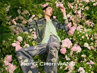 An East Asian male model reclines in a dense bed of pink and white flowers wearing a printed denim jacket and trousers for Feng Chen Wang.