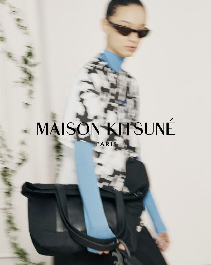 Model in sunglasses wearing a patterned top over a blue turtleneck and carrying a black shoulder bag against a white wall.