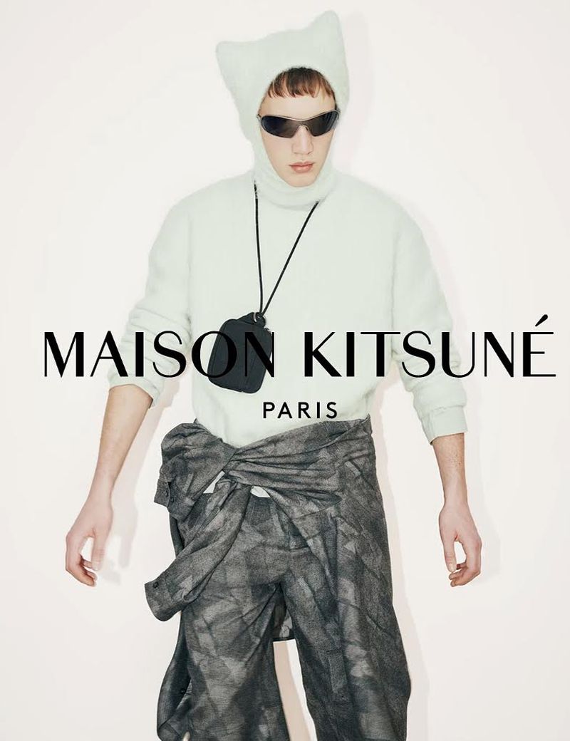 Model Achille Genet wearing a pale knit ear-hood balaclava, light sweater, and patterned wrap trousers for Maison Kitsuné FW26.