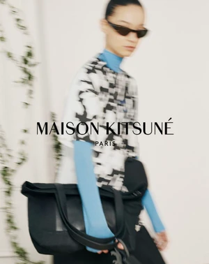 Model in sunglasses wearing a patterned top over a blue turtleneck and carrying a black shoulder bag against a white wall.