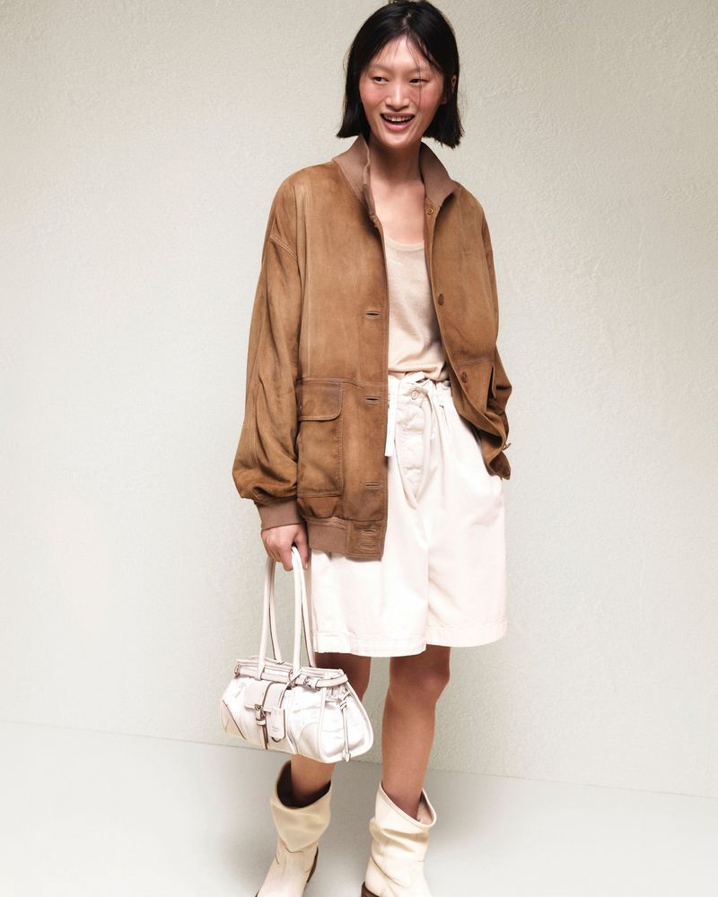 Model Yilan Hua smiling in a brown suede jacket, white shorts, cream boots, and carrying a white handbag against a pale backdrop.