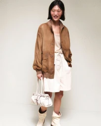 Model Yilan Hua smiling in a brown suede jacket, white shorts, cream boots, and carrying a white handbag against a pale backdrop.