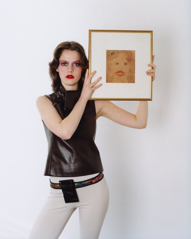 A woman in a sleeveless dark brown leather top holds a framed portrait sketch against a plain white studio backdrop.