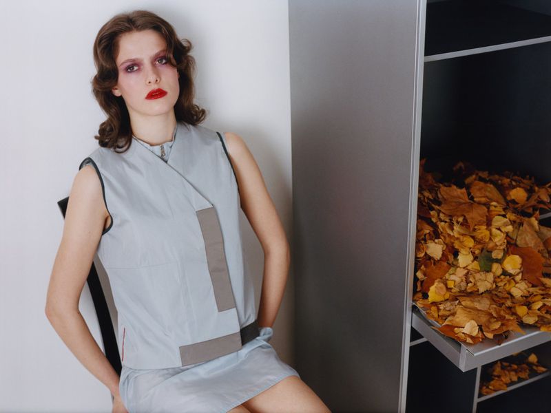 Fashion model seated beside a metal cabinet filled with dried autumn leaves against a white background.