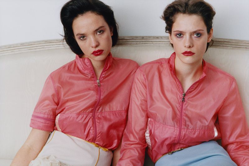 Two women wearing matching pink zip-up jackets seated side by side on a neutral upholstered sofa.
