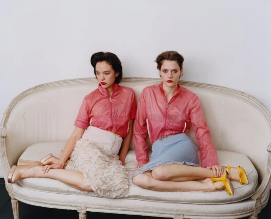Two women wearing translucent pink jackets sit on a vintage off-white sofa against a white backdrop.