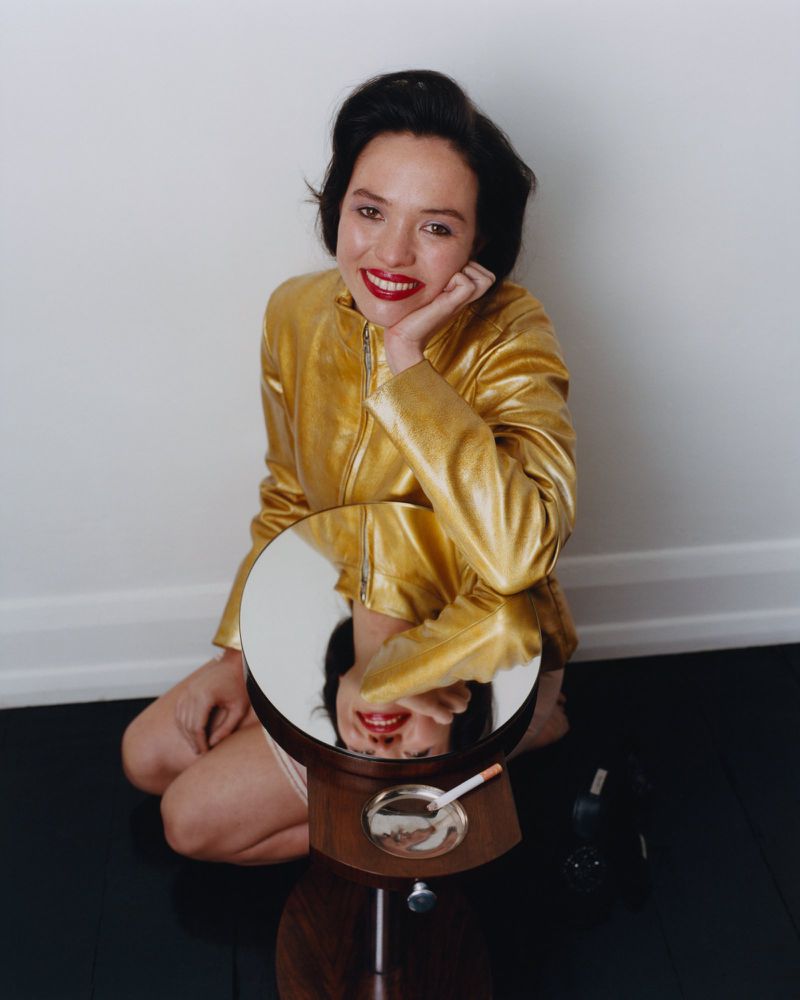 Woman in a metallic gold jacket smiling while kneeling beside a vanity mirror with an ashtray and cigarette.