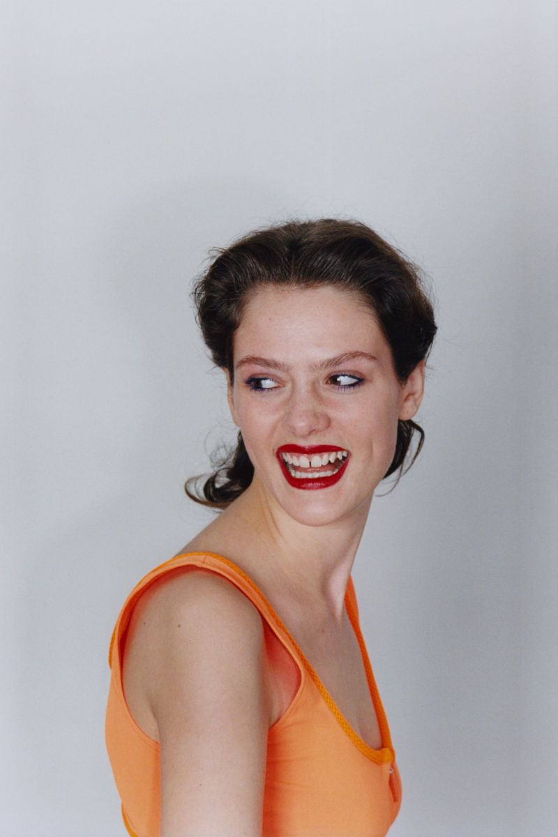 Woman with dark hair in an orange sleeveless top smiling over her shoulder against a white backdrop.