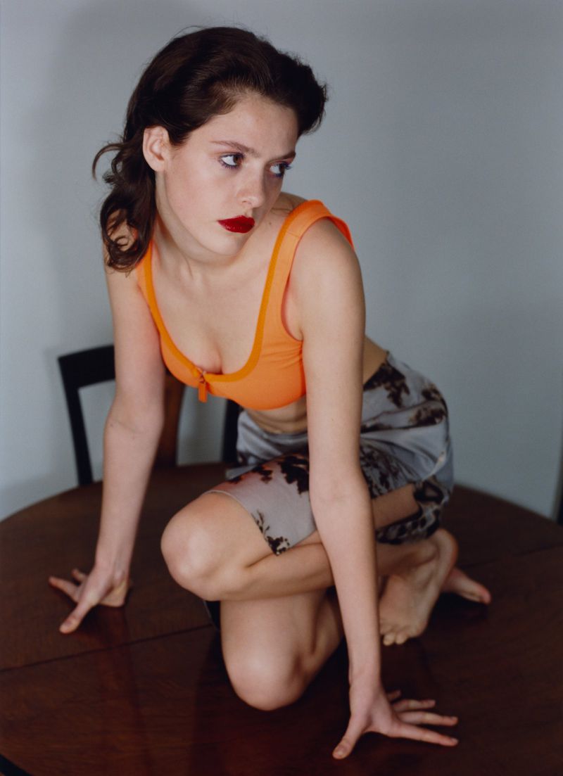 Young woman with bold red lipstick wearing an orange crop top and patterned shorts, kneeling barefoot on a polished wooden table.