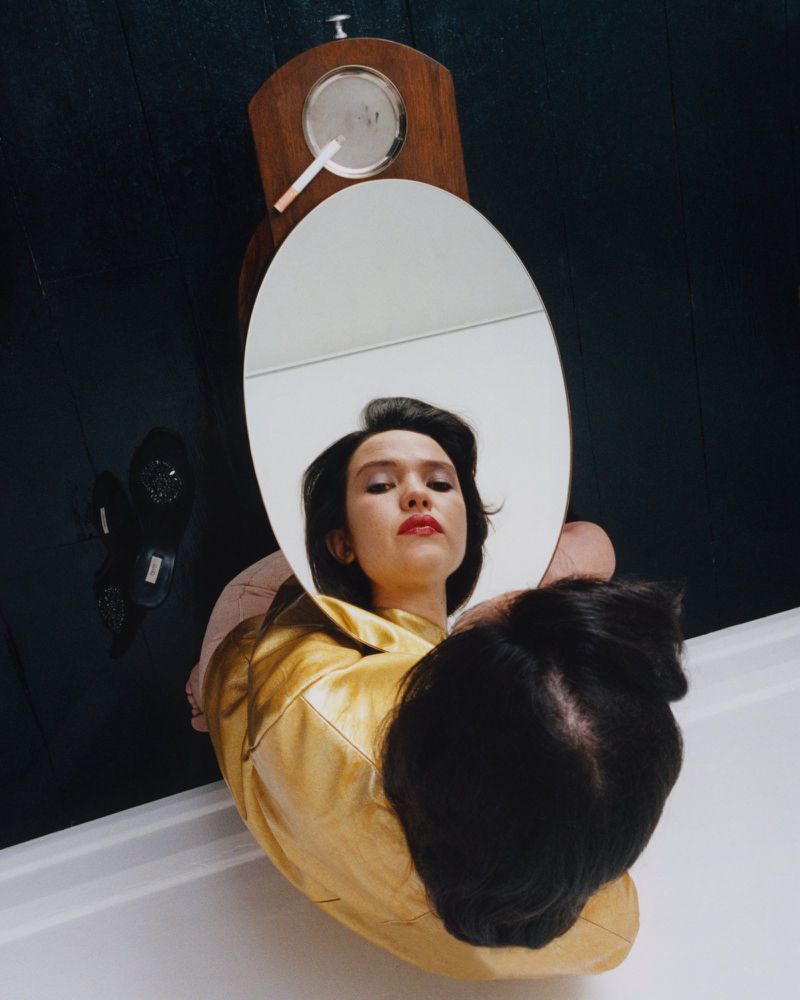 High-angle view of a woman in a gold jacket looking into an oval mirror showing her reflection with red lipstick.