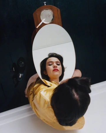 High-angle view of a woman in a gold jacket looking into an oval mirror showing her reflection with red lipstick.