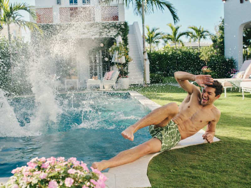A smiling shirtless man in green patterned swim trunks lounges by a swimming pool while getting splashed with water on a sunny lawn.