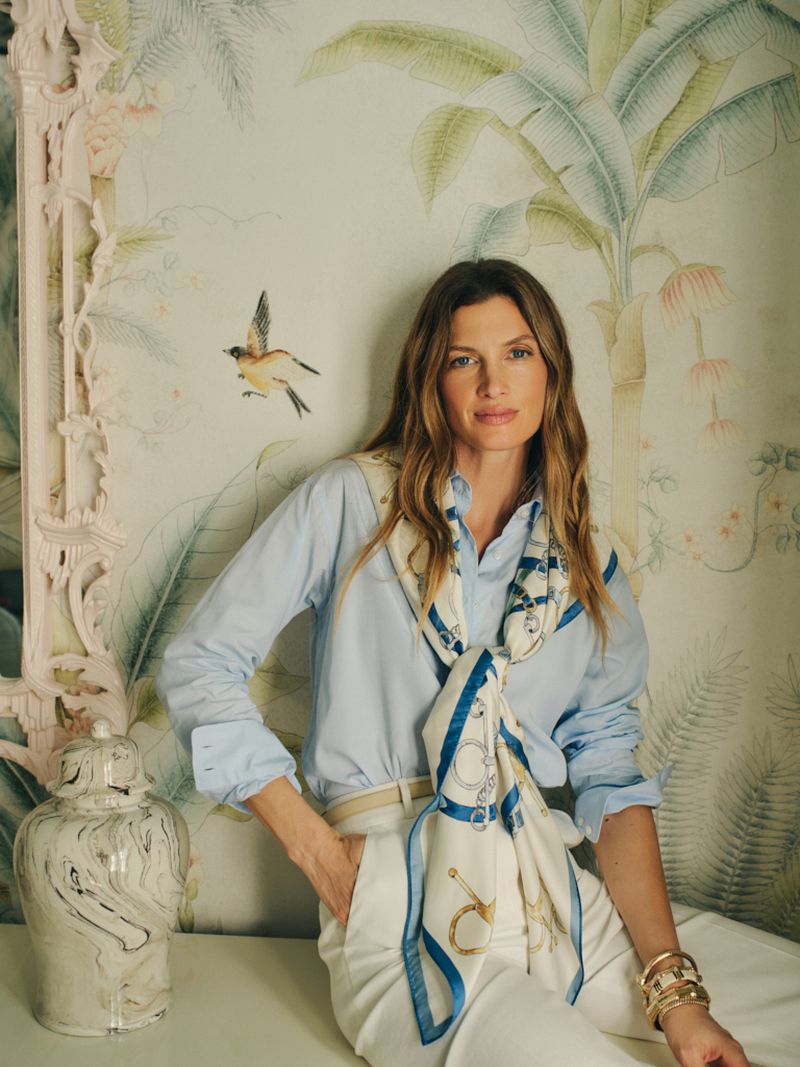 Woman wearing a light blue button-down shirt, printed silk scarf, and white trousers sitting against botanical wallpaper.