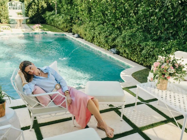Woman wearing a light blue button-up shirt and pink midi skirt reclining on a white patio armchair beside a swimming pool.