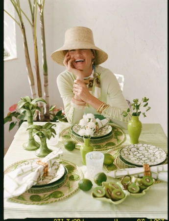 Woman wearing a wide-brim straw hat and light green top laughing behind a decorated green and white dining table.