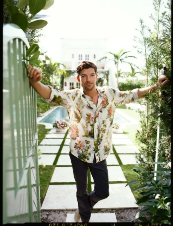 A man in a botanical print button-down shirt and dark trousers leaning against a garden gate in front of a pool.