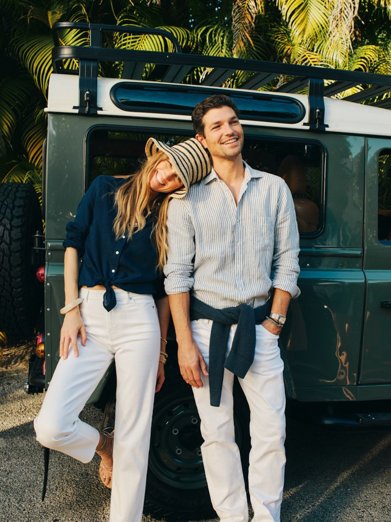 Man and woman smiling together in resortwear leaning against a vintage dark green off-road vehicle with palm trees behind.